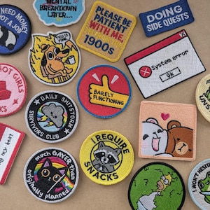 May include: A collection of embroidered patches featuring various designs including a dog in fire, a system error window, a raccoon with the text I REQUIRE SNACKS, a cat with a rainbow, a storm cloud, and other humorous phrases on a tan surface.