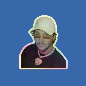 May include: A close-up of a man wearing a white baseball cap, sunglasses, and a black shirt with a large diamond heart pendant necklace.