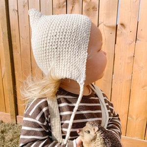 May include: A child wearing a white crocheted bonnet and a brown and white striped shirt holds a brown hedgehog figurine.