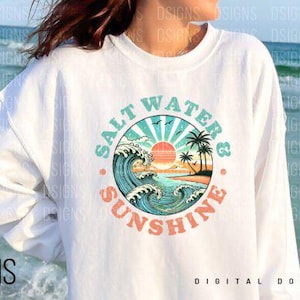 Pode incluir: Uma camisa de frio branca com um design gr&aacute;fico de um p&ocirc;r do sol sobre uma praia com ondas e palmeiras. O gr&aacute;fico est&aacute; em um c&iacute;rculo com o texto "Salt Water & Sunshine" ao redor.