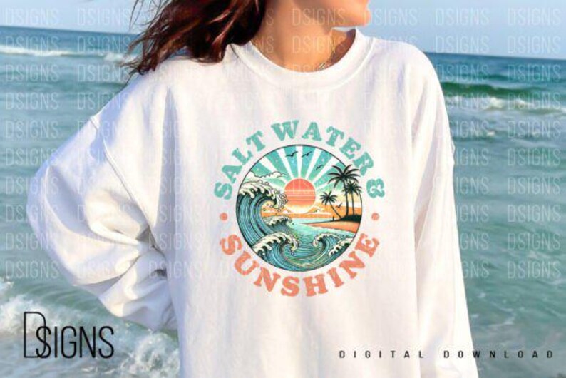 Pode incluir: Uma camisa de frio branca com um design gr&aacute;fico de um p&ocirc;r do sol sobre uma praia com ondas e palmeiras. O gr&aacute;fico est&aacute; em um c&iacute;rculo com o texto "Salt Water & Sunshine" ao redor.
