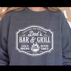 Puede incluir: Sudadera gris oscuro con un diseño blanco que dice "Dad's Bar & Grill" en un estilo vintage. El diseño incluye las frases "Chilling and Grilling", "Cold Brews", y "Good Times", junto con una ilustración de una parrilla.