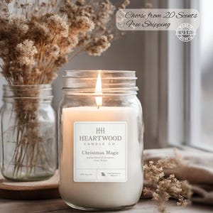 May include: A lit jar candle with a label that reads "Christmas Magic" by Heartwood Candle Co. The candle is in a clear jar, with a lit wick. The background includes dried flowers in a glass jar and a window.