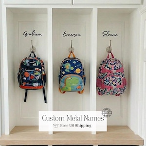 May include: Three children's backpacks hanging on hooks in a white cubby. The backpacks feature colorful designs, including a basketball theme, a dinosaur, and a floral pattern. The names "Graham", "Emerson", and "Sloane" are above each backpack. Text on the image reads "Custom Metal Names".