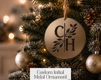 Puede incluir: Un adorno circular de metal en tono dorado que cuelga de un cordel, con un diseño recortado de las letras C y H separadas por una línea vertical, con motivos de hojas decorativas integrados en las letras en un árbol de Navidad.