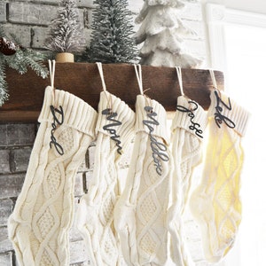 May include: Five white knitted stockings with personalized wooden name tags hanging on a wooden mantel. The names on the tags are "Dad", "Mom", "Mallory", "Joslyn", and "Jake".