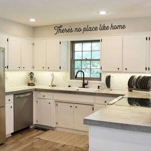 May include: A white kitchen with white cabinets, a stainless steel refrigerator, a stainless steel dishwasher, a white farmhouse sink, and a gray countertop. The kitchen has a window with a view of trees and a sign above the window that says "There's no place like home".