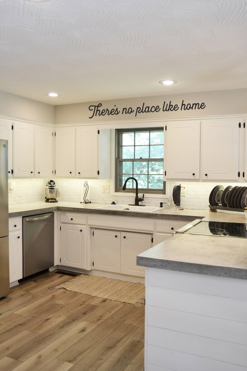 May include: A white kitchen with white cabinets, a stainless steel refrigerator, a stainless steel dishwasher, a white farmhouse sink, and a gray countertop. The kitchen has a window with a view of trees and a sign above the window that says "There's no place like home".