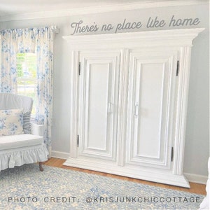 May include: White painted double door cabinet with a decorative top and the text "There's no place like home" above it. The cabinet is in a room with white walls, a window with blue and white floral curtains, and a white armchair with a blue and white floral pillow.