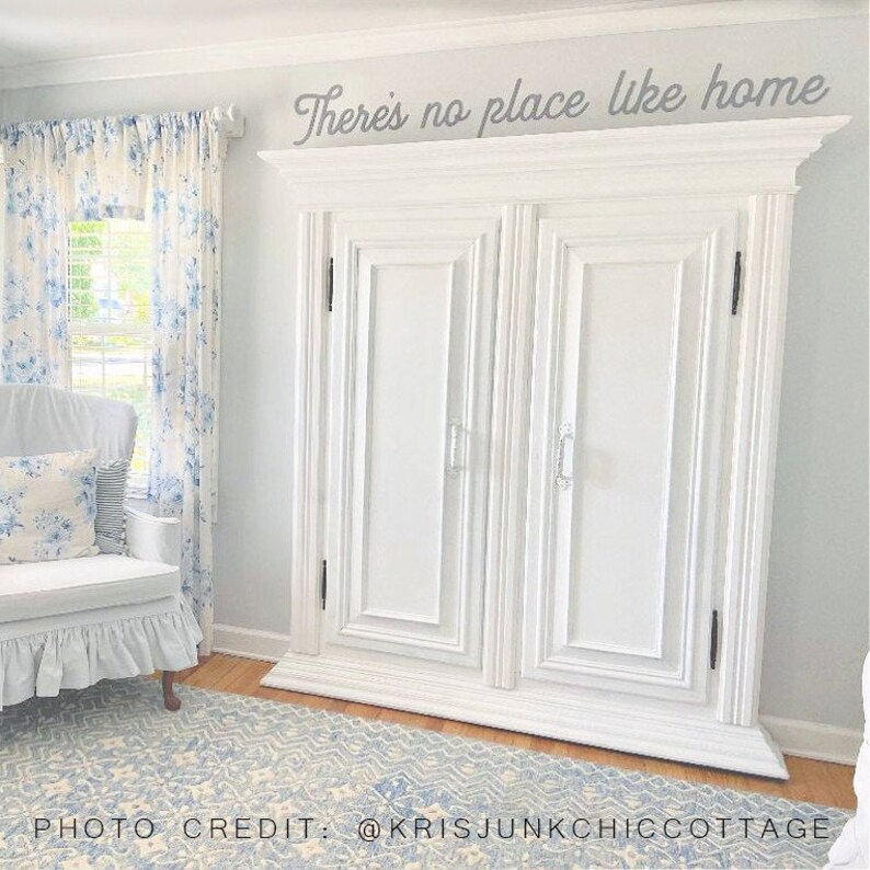 May include: White painted double door cabinet with a decorative top and the text "There's no place like home" above it. The cabinet is in a room with white walls, a window with blue and white floral curtains, and a white armchair with a blue and white floral pillow.