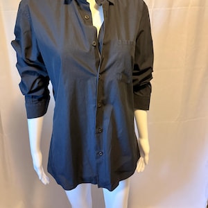 Pode incluir: Uma camisa azul marinho escuro com botões, gola clássica e um bolso no peito. A camisa de manga comprida é exibida em um manequim. A camisa está desabotoada na parte superior.