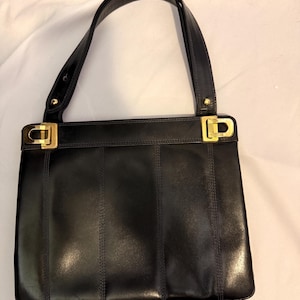 May include: A black leather handbag featuring a structured rectangular body with vertical stitched panel detailing. The bag includes a single shoulder strap attached with gold-tone metal hardware and matching gold-tone rectangular accents on the top corners.