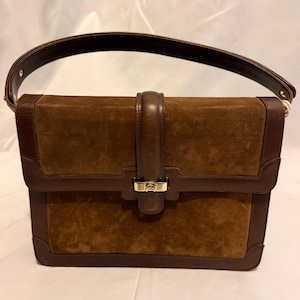 May include: A structured handbag featuring a brown suede body with dark brown leather trim and a single top handle. The bag includes a central vertical leather strap that secures with a gold-tone metal clasp featuring a small logo emblem.