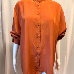 May include: A burnt orange button-down shirt with a mandarin collar, long sleeves with gathered cuffs, and a front placket featuring matching fabric-covered buttons. The garment has a smooth, slightly lustrous finish and a relaxed, loose fit.