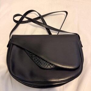 May include: A black shoulder bag with a curved flap closure. The bag features a smooth black leather body and a distinct crescent-shaped panel with a textured, scale-like pattern on the front flap. It includes a thin, adjustable shoulder strap.