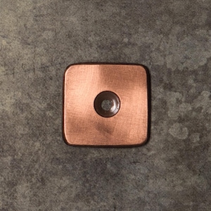 May include: A square-shaped copper-toned metal plate with rounded corners and a single centered circular hole. The surface features a brushed texture with visible linear markings across the metallic finish, creating a warm, reflective appearance.