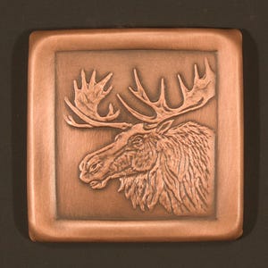 May include: A square, copper-colored relief carving of a moose head with large antlers. The moose is centered within a recessed square frame. The carving has detailed textures and is set against a dark background.