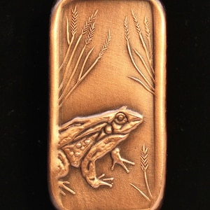 May include: A copper-colored metal bar with a raised design of a frog sitting in tall grass. The frog is facing the right side of the bar.