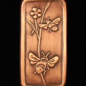 May include: A copper rectangle with a raised design of two bees and a flower. The bees are in flight and the flower has five petals.