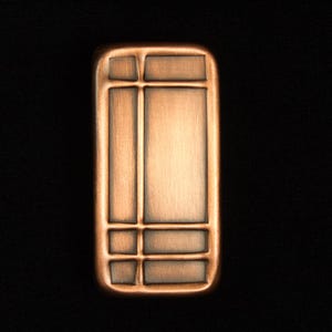 May include: A rectangular, copper-colored metal object with a geometric design. The design features a large central rectangle, two vertical rectangles, and four smaller squares. The object has rounded edges and is set against a black background.