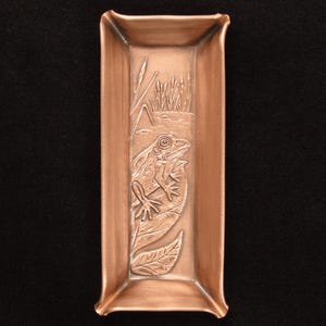 May include: A rectangular copper-colored tray with raised edges and a relief design featuring a frog sitting on a large leaf, surrounded by tall reeds and grasses. The metal surface has a brushed finish with darker accents highlighting the details.