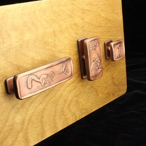 May include: Four copper cabinet pulls with nature-inspired designs. The pulls feature a squirrel, a fox, a bird, and a floral design. The pulls are mounted on a wooden surface.