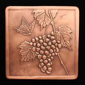 May include: A copper wall plaque with a raised design of a grapevine with a butterfly. The grapes are clustered together and the leaves are detailed. The plaque is signed "Vical" in the lower right corner.
