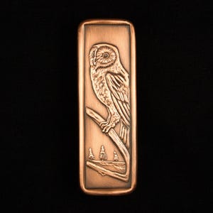 May include: A rectangular copper-colored metal object with a detailed relief carving of an owl perched on a branch. The owl is facing left, with a forest scene at the bottom. The background is black.