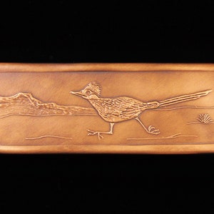 May include: A rectangular copper plaque with an embossed image of a roadrunner bird running across a desert landscape. The bird is facing right and has a long tail. The background shows a mountain range in the distance.