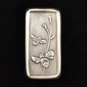 May include: A rectangular, silver-toned metal object with rounded edges. The surface features an embossed design of two bees and a flowering plant. The bees are detailed with wings and body segments. The background is a brushed metal finish.