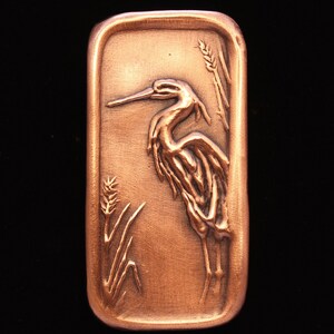May include: A copper-colored rectangular plaque with a raised image of a heron standing in reeds. The heron is facing left and has a long, thin beak. The reeds are on the left and right sides of the heron.