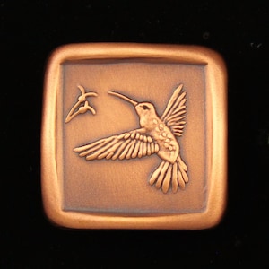 May include: A copper-colored square plaque with a raised image of a hummingbird in flight, with its beak extended towards a flower.