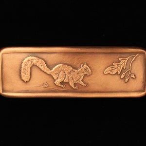 May include: A copper-colored rectangular plaque with an engraved image of a squirrel running to the right, with an acorn branch to the right of the squirrel.