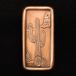 May include: A rectangular copper-colored metal object with a detailed relief of a desert scene. The design features a tall saguaro cactus, smaller desert plants, and a rock formation. The edges are slightly rounded, and the background is black.