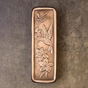 May include: A rectangular bronze-toned metal plate featuring a raised relief design of a hummingbird in flight surrounded by stylized flowers and leafy stems. The surface has a brushed metallic finish with darker recessed areas to highlight the details.