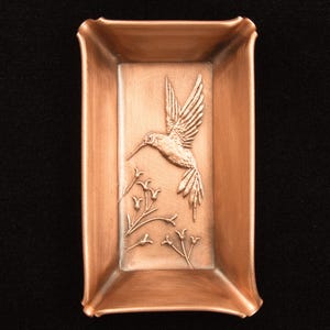 May include: A rectangular copper-colored tray featuring a raised relief design of a hummingbird in flight positioned above a cluster of small, delicate flowers. The tray has slightly curved edges and a smooth, metallic finish throughout the surface.