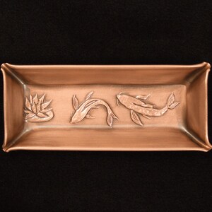 May include: A rectangular copper tray featuring a raised relief design on its interior base. The embossed artwork depicts a single lotus flower on the left side, followed by two swimming koi fish positioned horizontally across the center.
