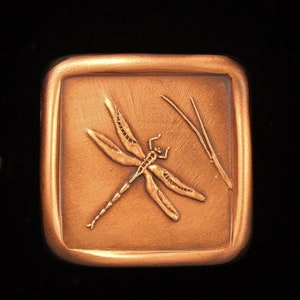 May include: A copper-colored square pendant with a raised image of a dragonfly and a stem. The pendant has a textured surface.