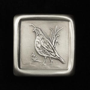 May include: A square, silver-toned metal belt buckle featuring a detailed relief of a bird, possibly a quail, standing near tall grass. The buckle has a rounded edge and a matte finish, set against a black background.