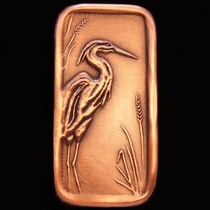 May include: A copper-colored metal plaque with a raised image of a heron standing in tall grass. The heron is facing right and has a long, thin beak. The grass is detailed with individual blades.