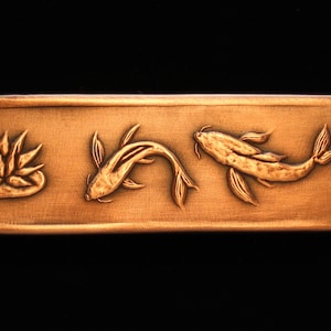 May include: A rectangular copper plaque with an engraved design of two koi fish and a lotus flower. The plaque has a warm, golden hue.