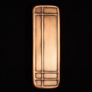 May include: A rectangular, copper-colored metal cabinet pull with a geometric design. The pull features a central vertical panel and horizontal and vertical lines creating a grid-like pattern. The edges are rounded, and the background is black.