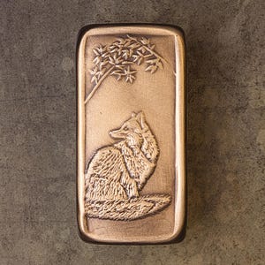 May include: A rectangular bronze-toned metal plaque featuring a raised relief design of a fox sitting beneath a leafy branch. The surface has a textured, weathered finish with darker recessed areas highlighting the detailed fur and foliage.