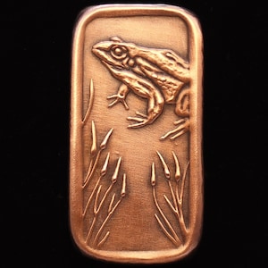 May include: A copper-colored metal rectangular plaque with a raised image of a frog sitting on a bed of reeds. The frog is facing the right side of the plaque.