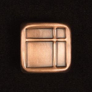 May include: A square, copper-colored cabinet knob with a grid design. The knob has a raised border and a textured surface, giving it a vintage or industrial aesthetic. The background is a dark, solid color.