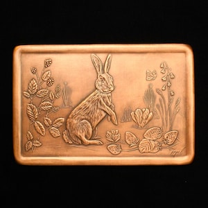 May include: A copper plaque with an embossed image of a rabbit in a field of flowers and berries. The plaque is signed "Yogel" in the lower right corner.
