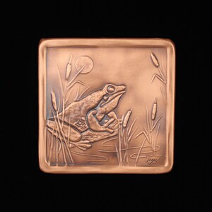 May include: A copper plaque with a raised relief image of a frog sitting on a log in a pond with cattails. The plaque is signed "Nigel".