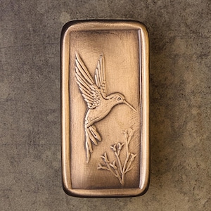 May include: A rectangular bronze-toned metal plaque featuring a raised relief design of a hummingbird in flight above a small plant. The surface has a brushed metallic finish with darker recessed areas that highlight the detailed bird and foliage.
