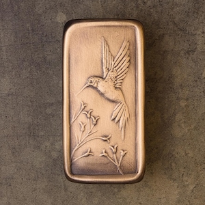 May include: A rectangular bronze-toned plaque featuring a raised relief design of a hummingbird in flight above a cluster of small, delicate flowers. The object has a smooth, metallic finish with darker shading in the recessed areas to highlight the details.