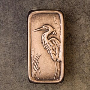 May include: A rectangular bronze-toned metal plaque featuring a raised relief design of a heron standing among tall reeds. The surface has a brushed, metallic finish with darker recessed areas that highlight the detailed bird and plant imagery.